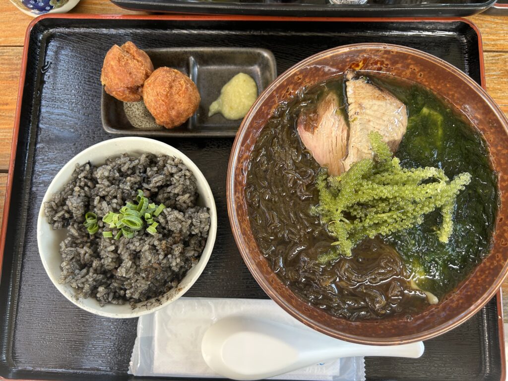 るま市「海をのむ」の沖縄そば。透明な魚介スープに海ぶどう、アーサ、もずくが乗り、三枚肉の代わりに厚切りのなまり節が添えられています